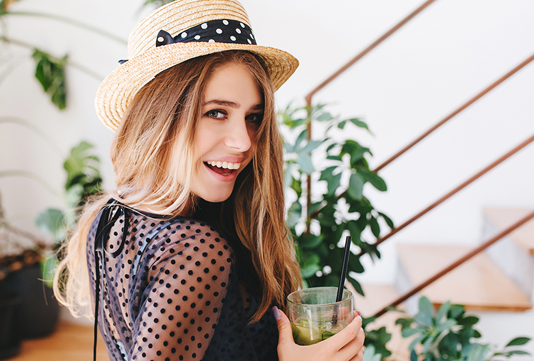 Inspired young woman with long shiny hair looking over shoulder and laughing holding glass of cold beverage. Indoor portrait of happy girl in hat with ribbon drink cocktail with plants on background