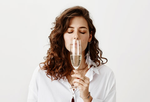 Studio portrait of beautiful sensual caucasian brunette smelling white wine while holding glass near nose, standing with closed eyes and enjoying taste over gray background